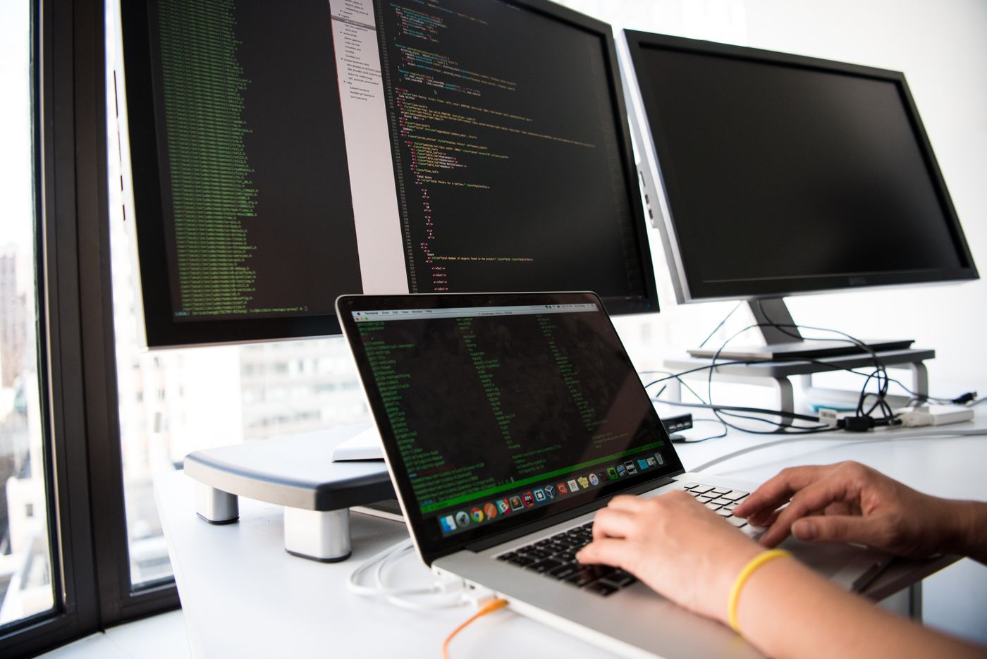A developer working at a laptop in front of two monitors showing code